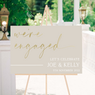 engagement party welcome sign with a cream background and gold lettering on a wooden easel stand