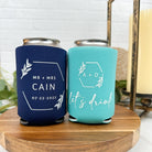 Wedding can coolers in the color of navy blue and turquoise, displayed on a round wooden platform with marble background and surrounding eucalyptus greenery.