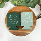 Wedding can coolers in the color of sage green and forest green, displayed on a round wooden platform with marble background and surrounding eucalyptus greenery.